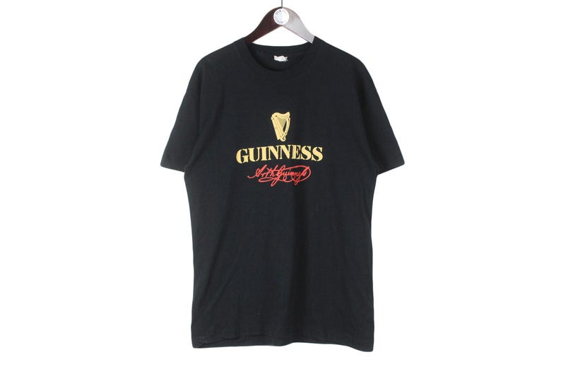 Puede incluir: Camiseta negra con el logo de Guinness, que incluye un arpa dorada y texto. La palabra "Guinness" es dorada, con la firma en rojo. La camiseta est&aacute; colgada en una percha de madera.
