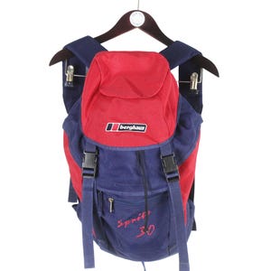 May include: A navy blue and red backpack featuring a top flap with a black and white Berghaus logo patch. The front includes two black plastic buckle closures, a zippered pocket, and the red embroidered text Sprite 30 on the lower section.