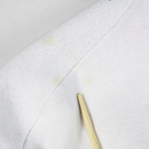 May include: Close-up of a white fabric, possibly a shirt or sweater, with visible yellow stains. A wooden stick is placed near the fabric. The texture of the material is visible, showing the weave of the threads.