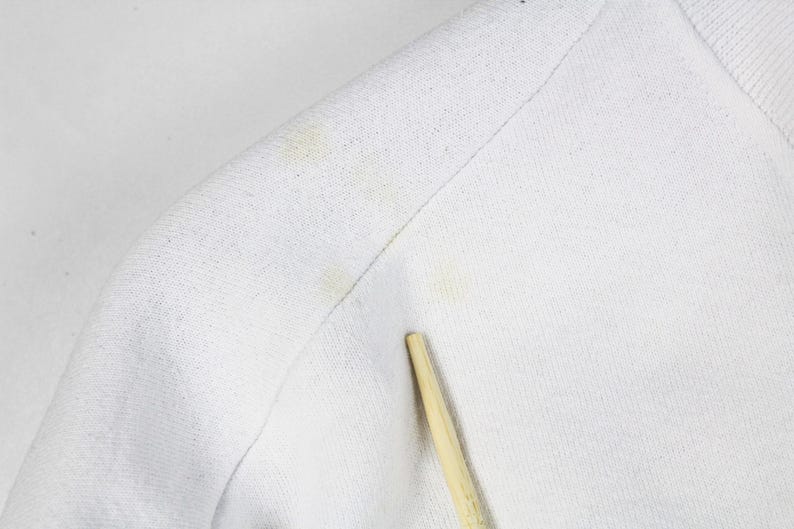 May include: Close-up of a white fabric, possibly a shirt or sweater, with visible yellow stains. A wooden stick is placed near the fabric. The texture of the material is visible, showing the weave of the threads.
