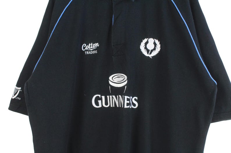 May include: Black rugby jersey with blue trim, featuring a white "Cotton Traders" logo, a white Guinness logo with a pint glass graphic, and a white thistle emblem.