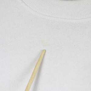 May include: Close-up of a white crew neck sweatshirt. The fabric has a textured appearance. A small stain is visible on the front. A wooden stick is placed in front of the sweatshirt.