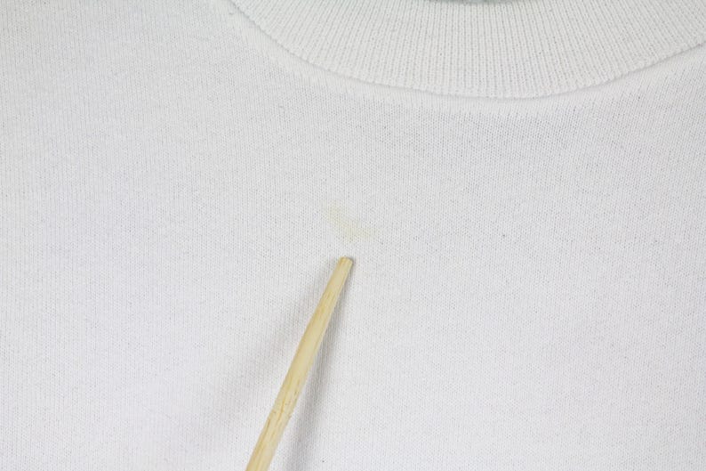 May include: Close-up of a white crew neck sweatshirt. The fabric has a textured appearance. A small stain is visible on the front. A wooden stick is placed in front of the sweatshirt.