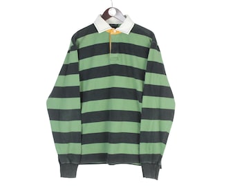 Puede incluir: Camiseta de rugby de manga larga con rayas horizontales verdes y gris carbón. La camiseta tiene cuello blanco, un detalle amarillo en el escote y puños de canalé. La camiseta está colgada de una percha de madera.