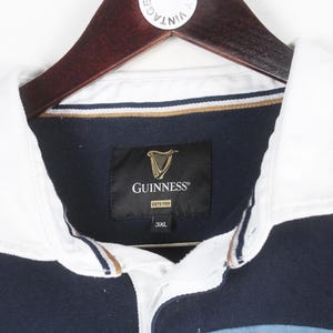 May include: A navy blue rugby shirt with a white collar and the Guinness logo. The shirt is hanging on a wooden hanger. The Guinness logo is a gold harp on a black label. The shirt is size 3XL.