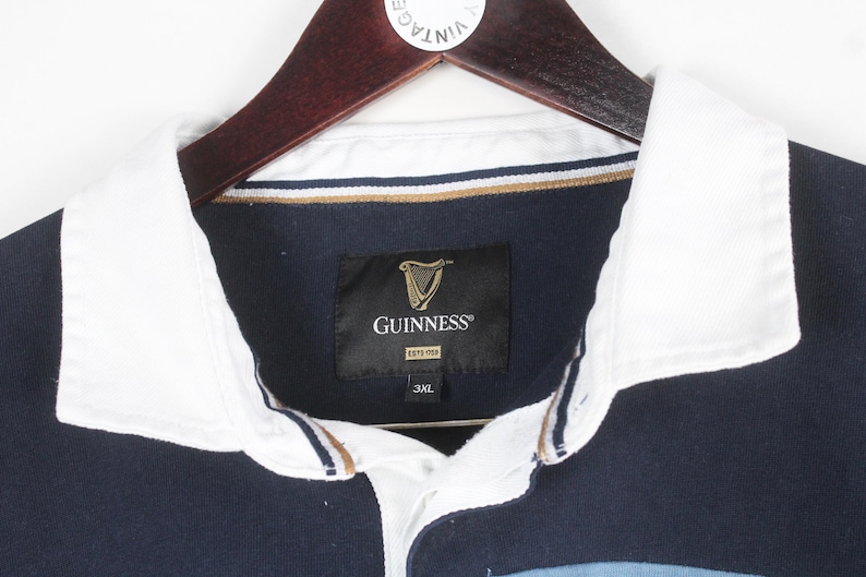 May include: A navy blue rugby shirt with a white collar and the Guinness logo. The shirt is hanging on a wooden hanger. The Guinness logo is a gold harp on a black label. The shirt is size 3XL.