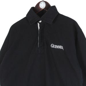May include: Black short-sleeved polo shirt with a white collar and a white placket. The word "Guinness" is embroidered in white on the chest. The shirt is hanging on a wooden hanger.