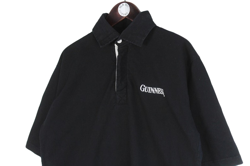 May include: Black short-sleeved polo shirt with a white collar and a white placket. The word "Guinness" is embroidered in white on the chest. The shirt is hanging on a wooden hanger.