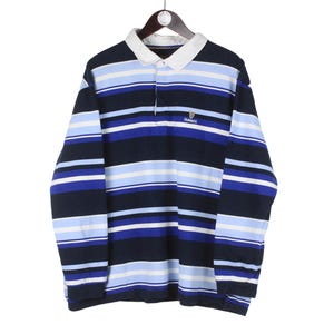 Puede incluir: Una camiseta de rugby de manga larga con rayas horizontales en azul marino, azul claro y blanco. La camiseta tiene un cuello blanco y un peque&ntilde;o logotipo en el pecho. La camiseta est&aacute; colgada en una percha de madera.