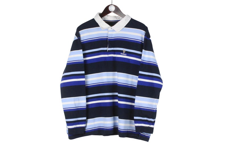 Puede incluir: Una camiseta de rugby de manga larga con rayas horizontales en azul marino, azul claro y blanco. La camiseta tiene un cuello blanco y un peque&ntilde;o logotipo en el pecho. La camiseta est&aacute; colgada en una percha de madera.