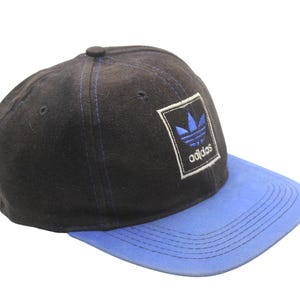 May include: Black and blue Adidas baseball cap. The cap has a black crown with blue stitching and a blue peak. The Adidas logo is on a white square patch on the front.