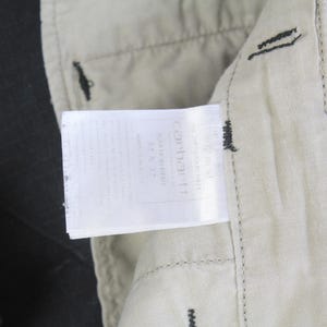Puede incluir: Primer plano de una etiqueta Carhartt adjunta a unos pantalones negros. La etiqueta blanca con texto negro est&aacute; cosida sobre una tela beige. Los pantalones son oscuros con costuras visibles.