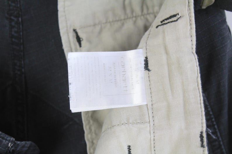 Puede incluir: Primer plano de una etiqueta Carhartt adjunta a unos pantalones negros. La etiqueta blanca con texto negro est&aacute; cosida sobre una tela beige. Los pantalones son oscuros con costuras visibles.