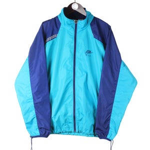 Op de afbeelding: Een blauwe en donkerblauwe windbreaker-jas met een ritssluiting en een Nike-logo op de borst. De jas heeft een capuchon met trekkoord en elastische manchetten.