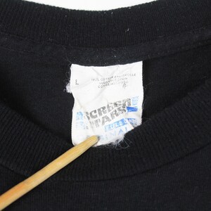 Puede incluir: Primer plano de un cuello de camiseta negra con una etiqueta blanca. La etiqueta tiene texto azul y negro, incluyendo la marca "Screen Stars" y la talla "L". La camiseta es de algod&oacute;n.