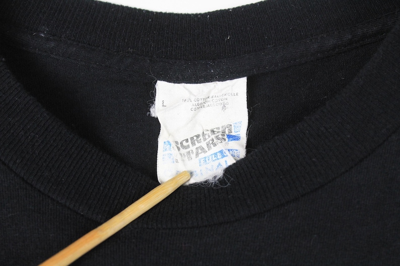 Puede incluir: Primer plano de un cuello de camiseta negra con una etiqueta blanca. La etiqueta tiene texto azul y negro, incluyendo la marca "Screen Stars" y la talla "L". La camiseta es de algod&oacute;n.