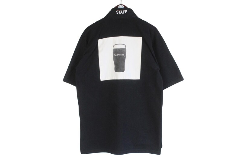 May include: Black short-sleeved shirt with a white square on the back featuring a Guinness pint glass graphic. The word "STAFF" is embroidered on the collar.