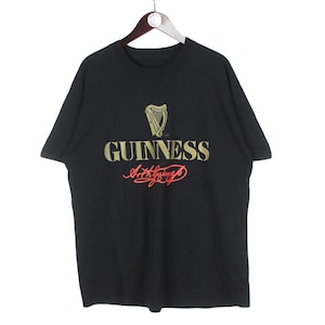 Op de afbeelding: Zwart t-shirt met het Guinness-logo in goud en rood. Het logo toont een harp en het woord "GUINNESS" boven een cursief handschrift. Het t-shirt hangt aan een houten hanger.