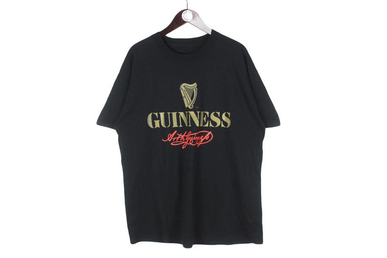 Op de afbeelding: Zwart t-shirt met het Guinness-logo in goud en rood. Het logo toont een harp en het woord "GUINNESS" boven een cursief handschrift. Het t-shirt hangt aan een houten hanger.