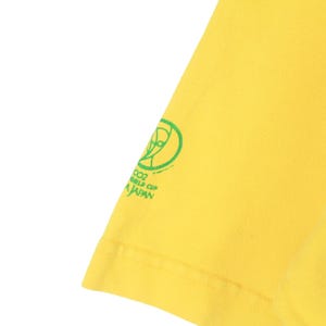 May include: Bright yellow t-shirt with a green graphic and text. The graphic includes a stylized globe and the text reads "2002 WORLD CUP JAPAN". The shirt's fabric is visible, showing its texture and color.