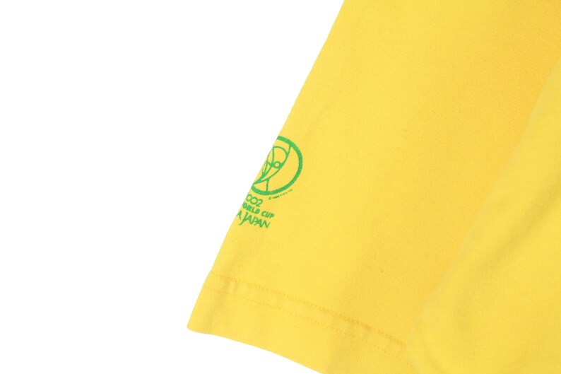 May include: Bright yellow t-shirt with a green graphic and text. The graphic includes a stylized globe and the text reads "2002 WORLD CUP JAPAN". The shirt's fabric is visible, showing its texture and color.