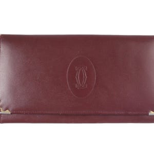 Może przedstawiać: Bordowy skórzany portfel o prostokątnym kształcie. Portfel posiada złote detale narożne i centralnie wytłoczone logo. Portfel jest zamknięty i wydaje się nowy.