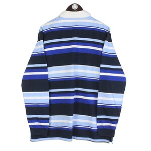 May include: A long-sleeved rugby shirt with horizontal stripes in shades of blue, white, and navy. The shirt has a white collar and cuffs. The garment is displayed on a hanger against a white background.