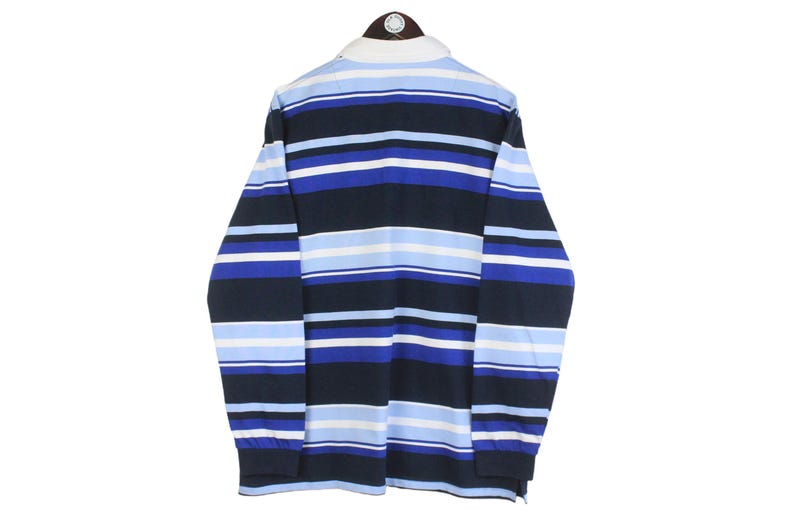 May include: A long-sleeved rugby shirt with horizontal stripes in shades of blue, white, and navy. The shirt has a white collar and cuffs. The garment is displayed on a hanger against a white background.