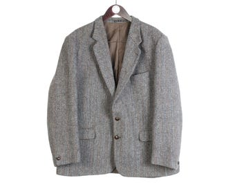 May include: A gray tweed blazer with a classic design, featuring a notched lapel, two front pockets, and three buttons. The jacket is hanging on a wooden hanger against a white background. The blazer's fabric has a subtle herringbone pattern.