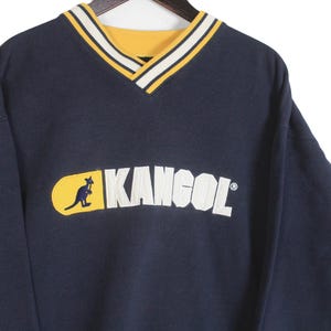K&ouml;nnte beinhalten: Marineblaues Kangol-Sweatshirt mit gelb-wei&szlig; gestreiftem V-Ausschnitt. Das Kangol-Logo mit einem K&auml;nguru ist wei&szlig; auf einem gelben Oval abgebildet.