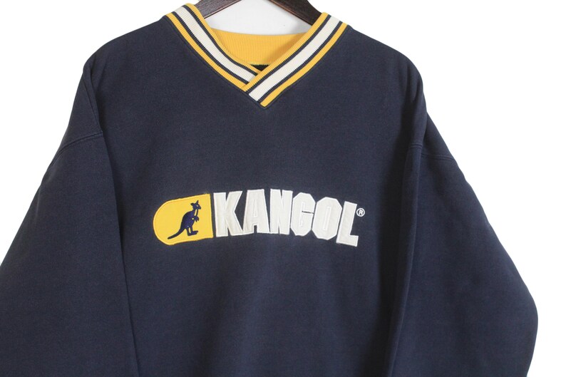 K&ouml;nnte beinhalten: Marineblaues Kangol-Sweatshirt mit gelb-wei&szlig; gestreiftem V-Ausschnitt. Das Kangol-Logo mit einem K&auml;nguru ist wei&szlig; auf einem gelben Oval abgebildet.