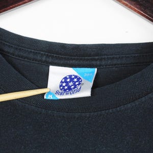 Pu&ograve; includere: Primo piano di una maglietta nera con un'etichetta. L'etichetta &egrave; bianca e blu con la scritta "Starworld" e un disegno di stelle bianche. Un puntatore di legno tocca l'etichetta.