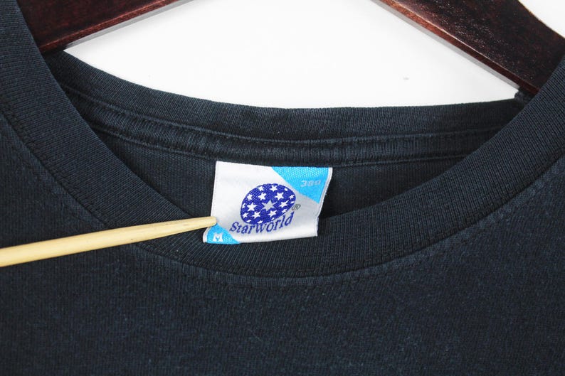 Pu&ograve; includere: Primo piano di una maglietta nera con un'etichetta. L'etichetta &egrave; bianca e blu con la scritta "Starworld" e un disegno di stelle bianche. Un puntatore di legno tocca l'etichetta.