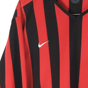 Puede incluir: Camiseta deportiva a rayas rojas y negras con cuello en V. La camiseta presenta un logotipo blanco de Nike en el pecho. La tela tiene un patr&oacute;n de rayas verticales.
