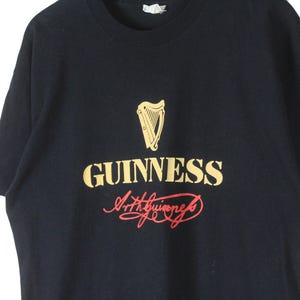Puede incluir: Camiseta negra con el logo de Guinness en dorado y rojo. El logo incluye un arpa sobre la palabra "GUINNESS" y la firma "Arthur Guinness". Una prenda de vestir cl&aacute;sica.