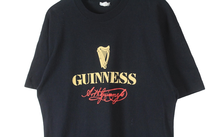 Puede incluir: Camiseta negra con el logo de Guinness en dorado y rojo. El logo incluye un arpa sobre la palabra "GUINNESS" y la firma "Arthur Guinness". Una prenda de vestir cl&aacute;sica.