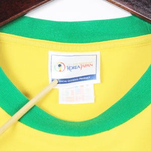 May include: Yellow t-shirt with a green collar and sleeve trim. The shirt has a tag that reads "KOREA JAPAN OFFICIAL LICENSED PRODUCT". The shirt is hanging on a wooden hanger.
