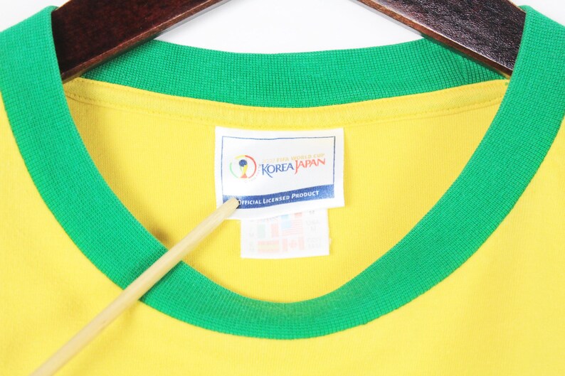 May include: Yellow t-shirt with a green collar and sleeve trim. The shirt has a tag that reads "KOREA JAPAN OFFICIAL LICENSED PRODUCT". The shirt is hanging on a wooden hanger.