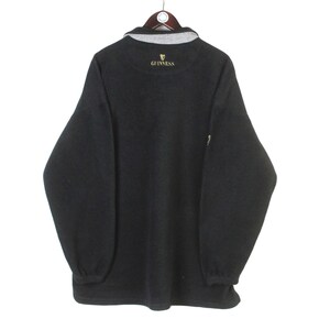 May include: Dark gray fleece pullover with a gray collar and the Guinness logo embroidered in gold. The long-sleeved shirt has a relaxed fit and ribbed cuffs. The garment is hanging on a wooden hanger.
