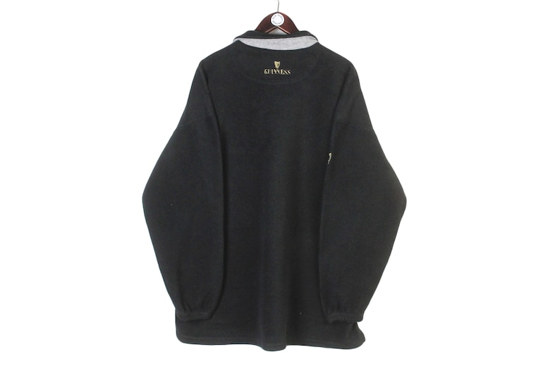 May include: Dark gray fleece pullover with a gray collar and the Guinness logo embroidered in gold. The long-sleeved shirt has a relaxed fit and ribbed cuffs. The garment is hanging on a wooden hanger.