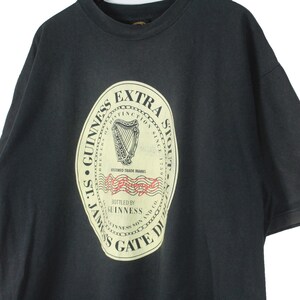 May include: Black t-shirt featuring a large, cream-colored Guinness Extra Stout label. The label includes a harp, text, and the words "St. James's Gate Dublin". A vintage style tee.