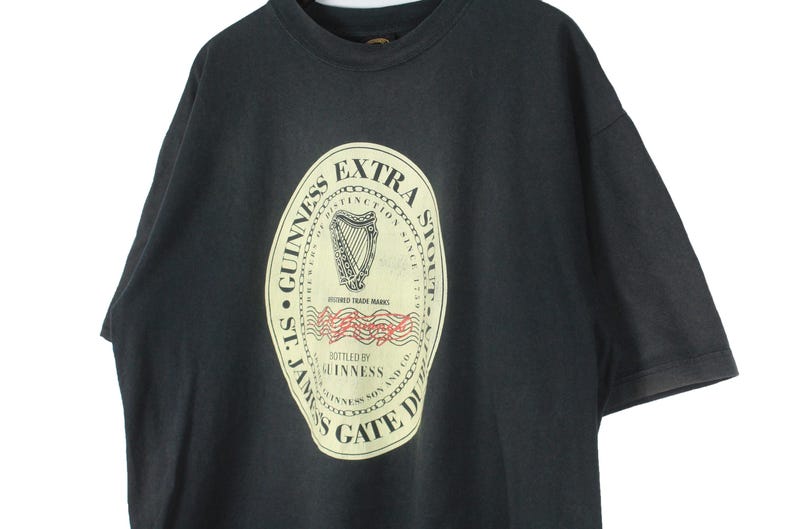 May include: Black t-shirt featuring a large, cream-colored Guinness Extra Stout label. The label includes a harp, text, and the words "St. James's Gate Dublin". A vintage style tee.