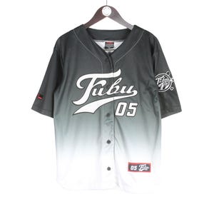 Può includere: Una maglia da baseball a maniche corte con bottoni e una sfumatura di colore dal nero al bianco. La parte anteriore mostra la scritta bianca Fubu e il numero 05. Un logo circolare con il testo Fubu 05 si trova sulla manica sinistra e una piccola toppa rettangolare è posizionata in basso.