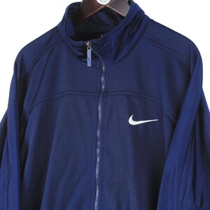May include: A navy blue Nike track jacket with a full zipper and a high collar. The jacket features a white Nike swoosh logo on the chest. The jacket is hanging on a wooden hanger.