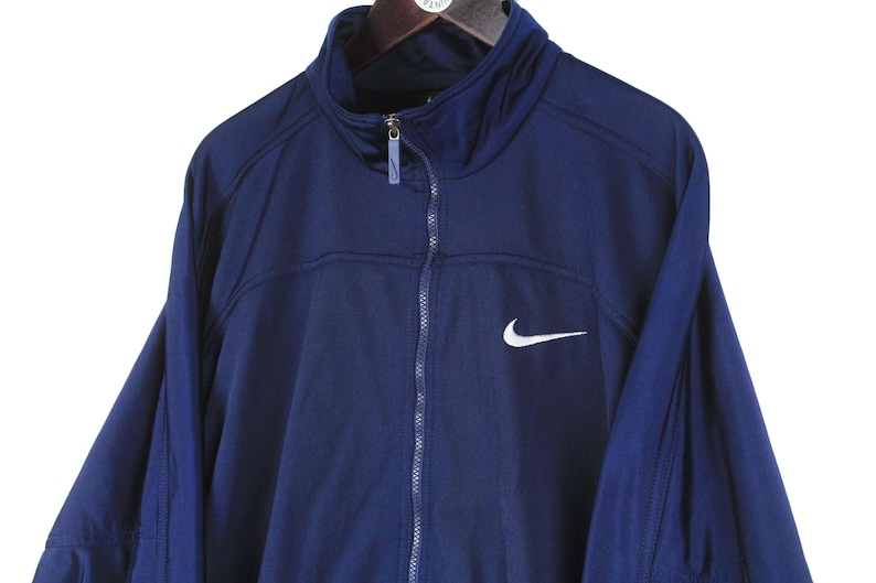 May include: A navy blue Nike track jacket with a full zipper and a high collar. The jacket features a white Nike swoosh logo on the chest. The jacket is hanging on a wooden hanger.