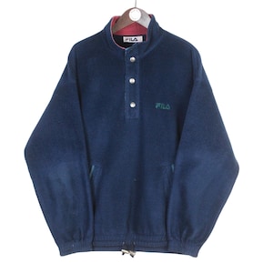 K&ouml;nnte beinhalten: Marineblaues FILA-Fleece-Pullover mit hohem Kragen und drei Kn&ouml;pfen. Der Kragen hat ein rotes Futter. Das FILA-Logo ist gr&uuml;n auf der Brust gestickt. Der untere Saum hat einen Kordelzug.