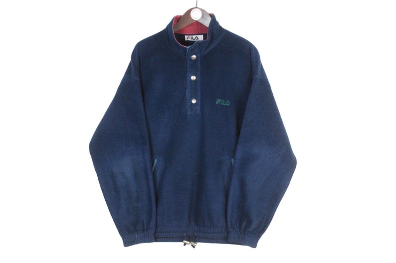 K&ouml;nnte beinhalten: Marineblaues FILA-Fleece-Pullover mit hohem Kragen und drei Kn&ouml;pfen. Der Kragen hat ein rotes Futter. Das FILA-Logo ist gr&uuml;n auf der Brust gestickt. Der untere Saum hat einen Kordelzug.