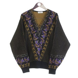 Puede incluir: Un suéter vintage con cuello en V y un patrón geométrico. El suéter presenta una base negra con un diseño central en morado, dorado y negro. Las mangas y el dobladillo inferior son de color negro liso, y cuelga de una percha de madera.