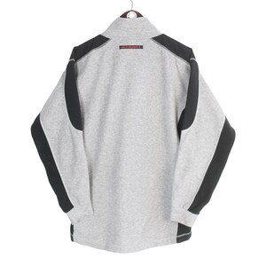 Pu&ograve; includere: Una felpa grigia con dettagli neri sulle spalle e sulle maniche. La felpa ha un collo alto e un piccolo dettaglio rosso vicino al colletto. Le maniche sono lunghe e i polsini sono a coste. La felpa &egrave; appesa a un'appendiabiti.