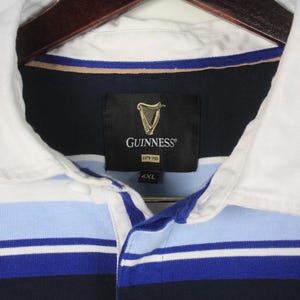 Puede incluir: Una camiseta de rugby a rayas azul marino, azul claro y blanco con cuello blanco. La camiseta presenta el logotipo de Guinness en una etiqueta negra. Talla 4XL.
